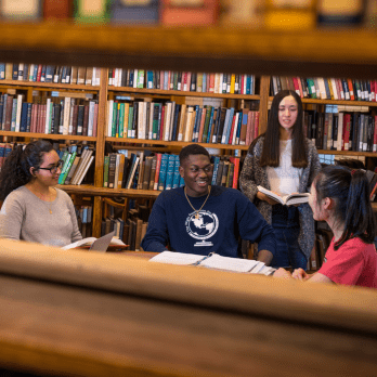 A group of four students sit and stand around a table in a library, studying and smiling, with shelves of books in the background.