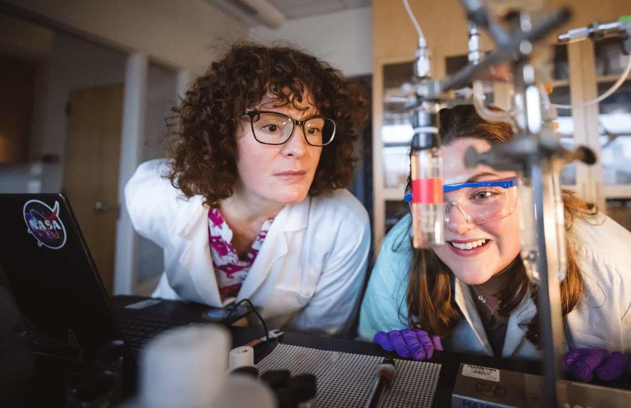 Two scientists wearing lab coats and safety glasses work together in a laboratory, closely observing an experiment with glassware and equipment, appearing focused and engaged.
