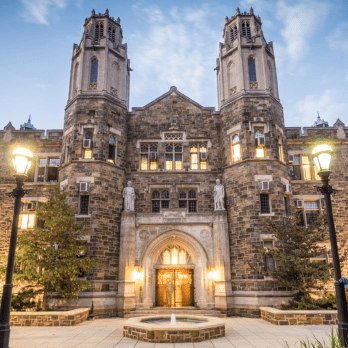 A grand stone building with two tall, ornate towers and arched entrance, illuminated by exterior lights at dusk, showcasing Gothic architectural details.