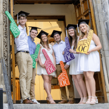Six graduates in caps and gowns stand on steps outside a building, smiling and holding colorful foam fingers. They are celebrating together in front of open wooden doors.