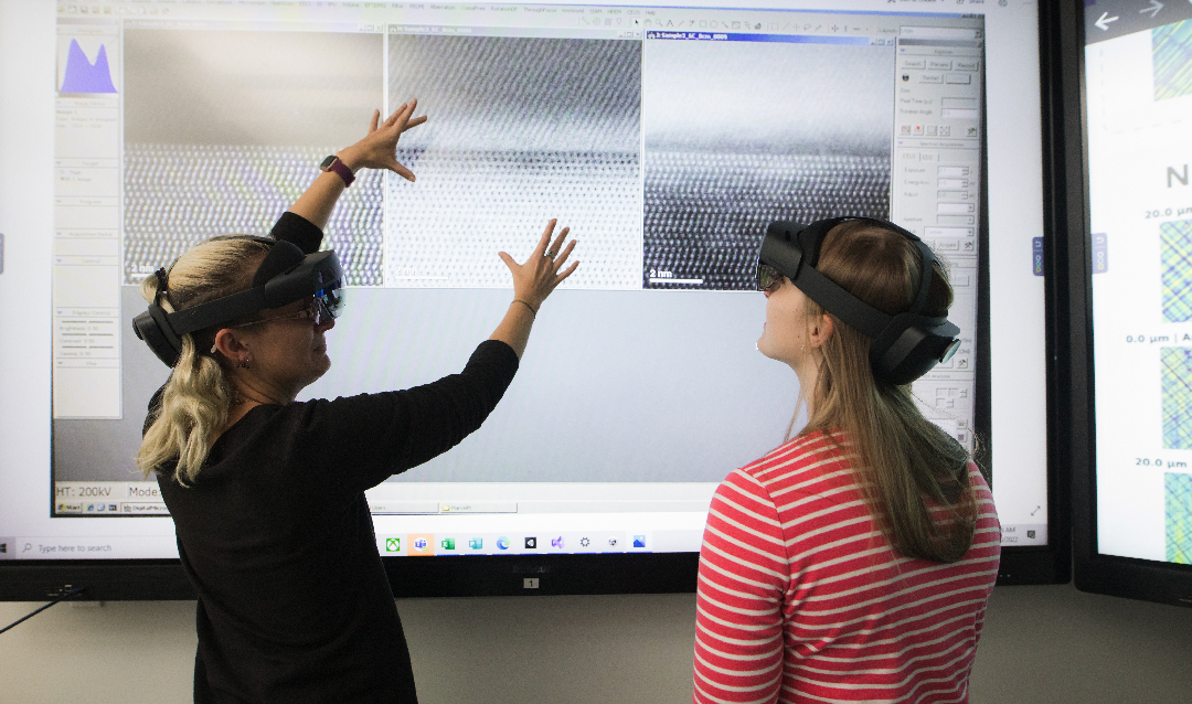 Two women wear VR headsets in front of a large touchscreen