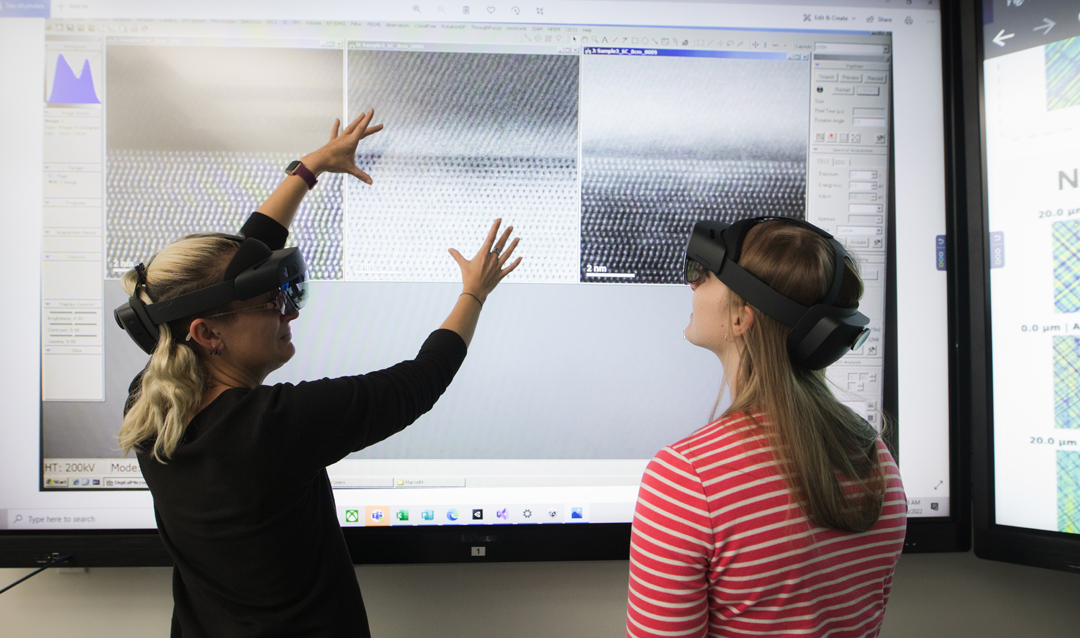 Two women wear VR headsets in front of a large touchscreen