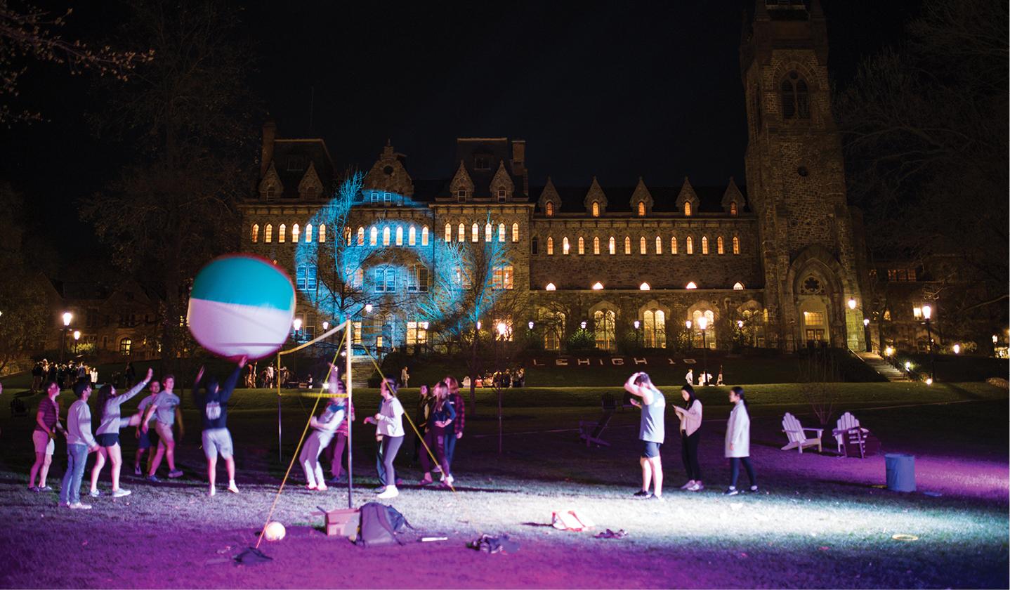 People are gathered in an open area at night. A large illuminated spherical object with blue and white swirls is in the foreground. A brightly lit, elaborate building with several towers and decorative lighting is visible in the background.