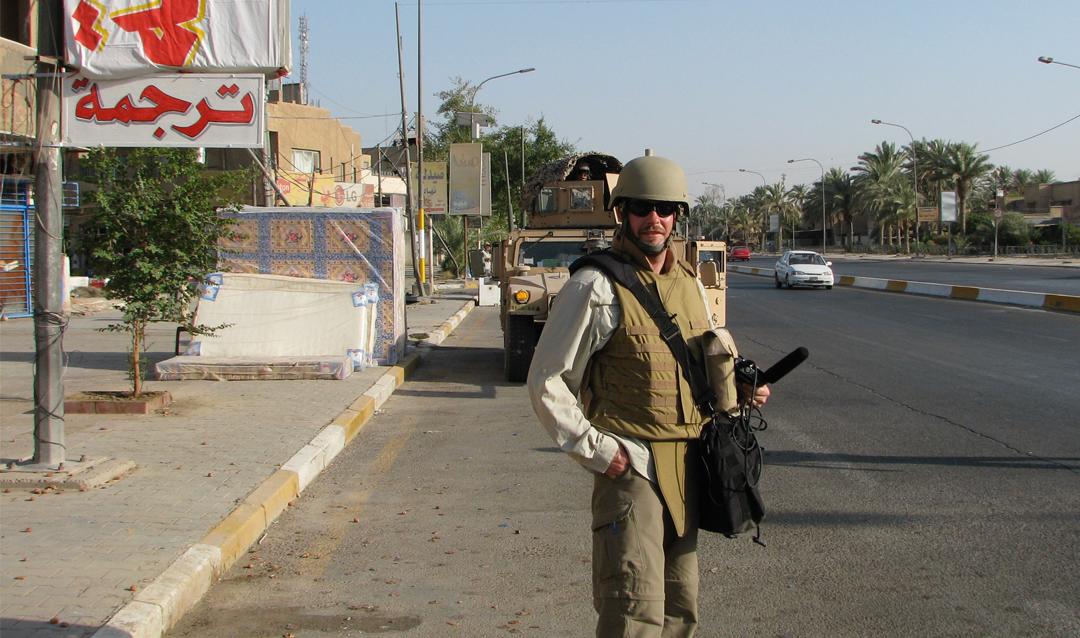 A Former War Correspondent, Sean Carberry, in military gear stands on a street with armored vehicles in the background.