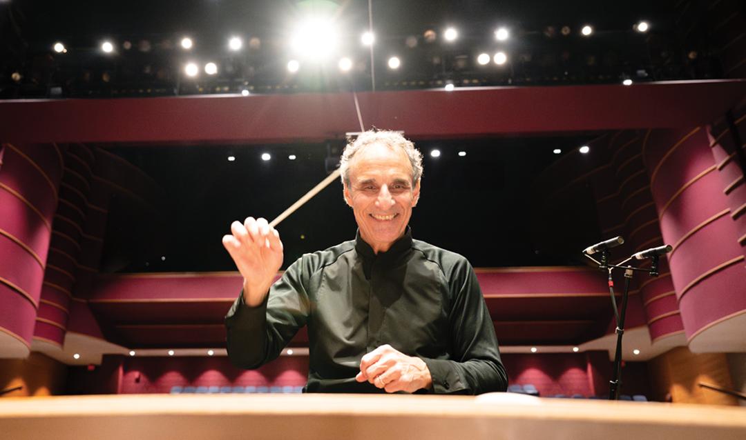 Conductor, Paul Salerni, with baton smiling at the camera from the stage of an empty auditorium showcasing Lehigh University's exquisite architecture.