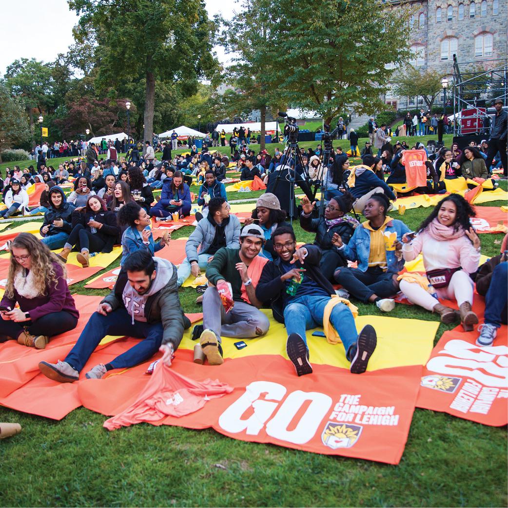 A large group of people sits on colorful blankets spread across a grassy area, enjoying an outdoor event