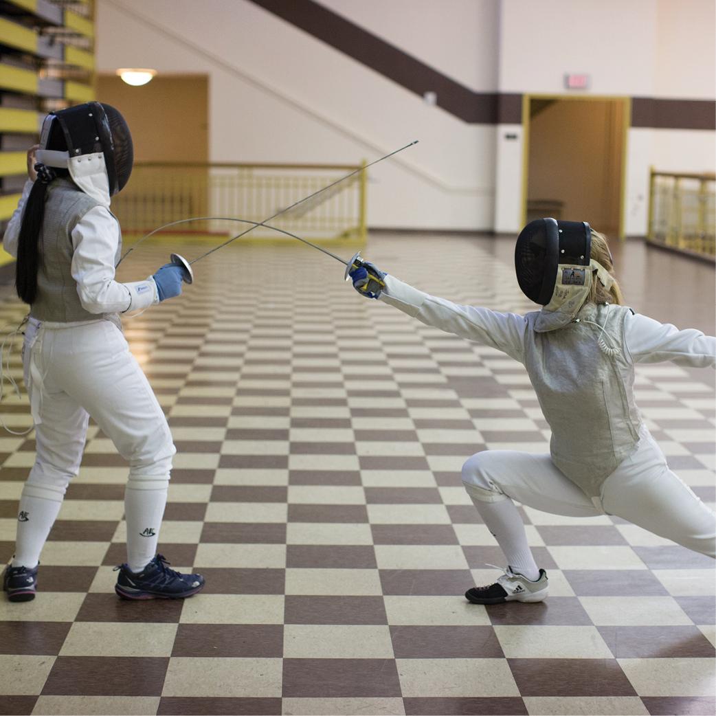 Two fencers dressed in white protective gear and black masks are engaging in a duel in an indoor hall with a checkered floor. One fencer is lunging forward with their weapon extended, while the other stands in a defensive stance.