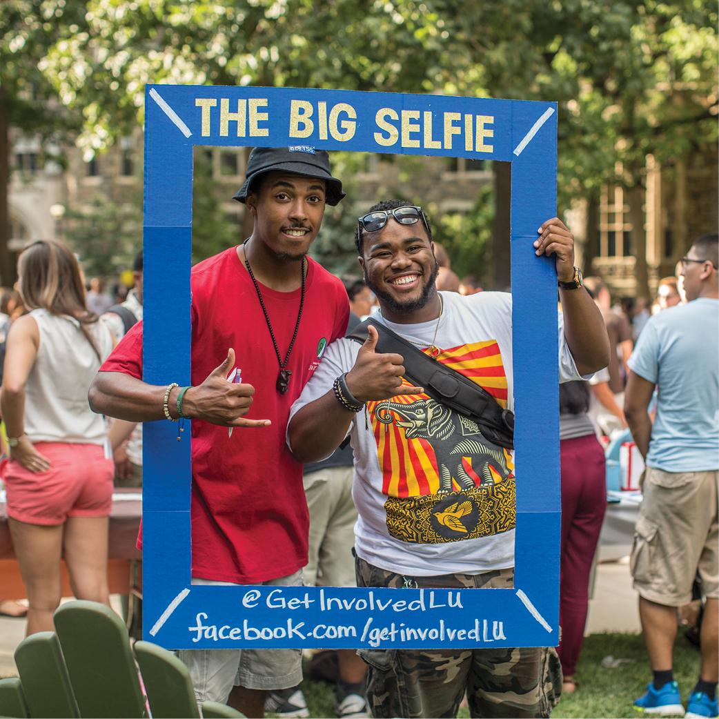 Two smiling men are posing with their thumbs up in a photo frame that reads "The Big Selfie." The bottom of the frame has a hashtag and web link.