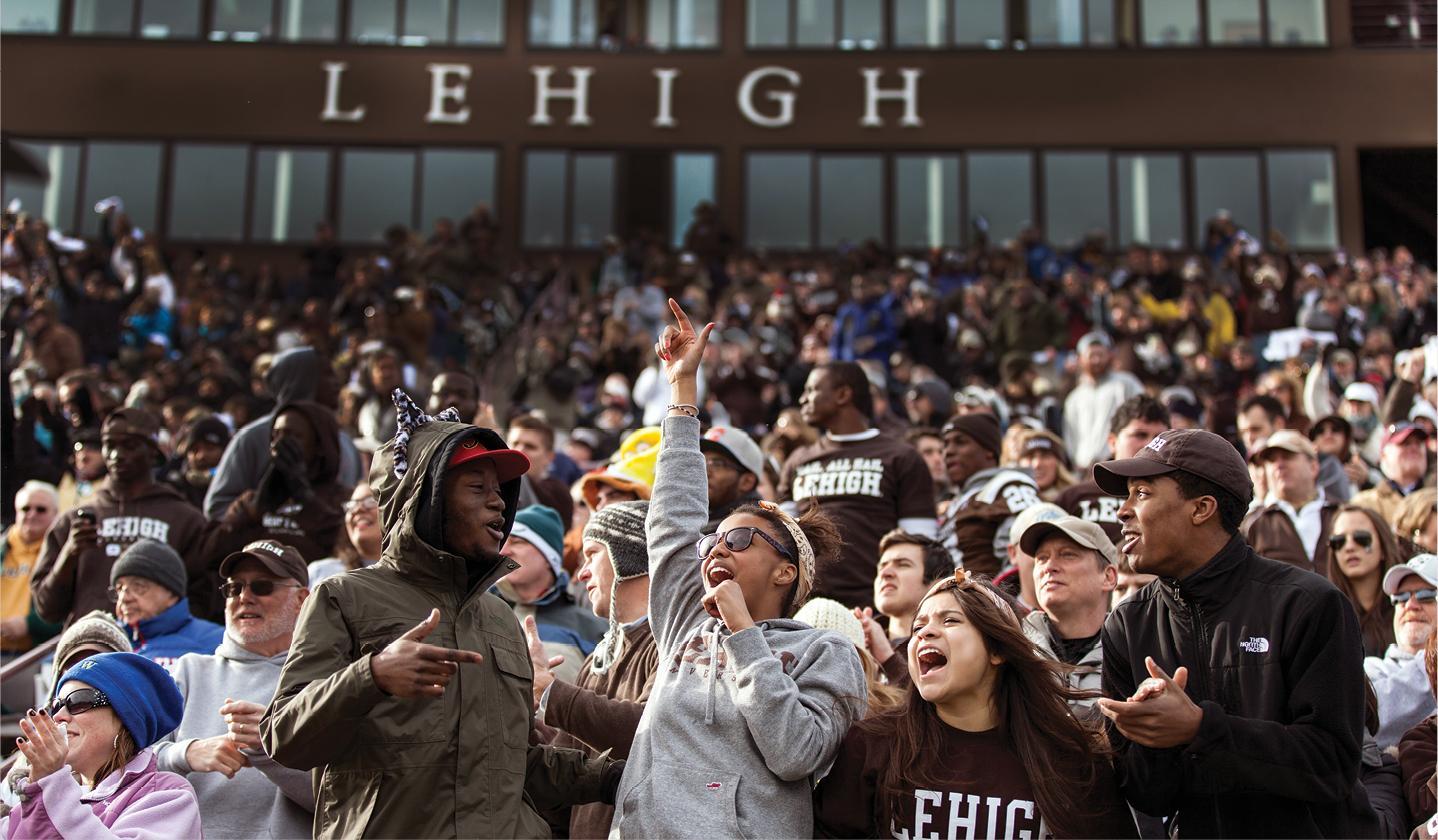 A lively crowd of people cheering and raising their hands in front of a building with large windows. The building has "LEHIGH" written across the top. 