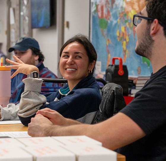 A group of people sit at a table, engaging in a cheerful conversation. A woman in the center smiles while a man to her right gestures as he speaks. In the background, there is a colorful world map on the wall and some electronic equipment on the table.