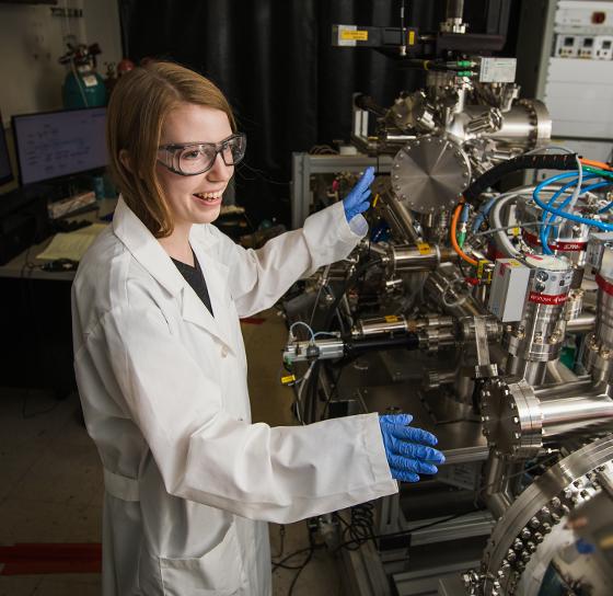 A person in a lab coat and safety glasses is working with complex scientific equipment. The individual is wearing blue gloves and standing in front of a machine with various knobs, wires, and metallic components. Behind them, a desk with more equipment is visible.