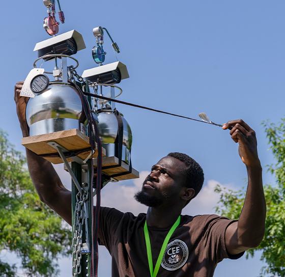 A person is adjusting equipment on a tall pole outdoors. They are holding a cable attached to devices, possibly for atmospheric data collection or environmental monitoring. The sky is clear and blue, and leafy green trees are visible in the background.