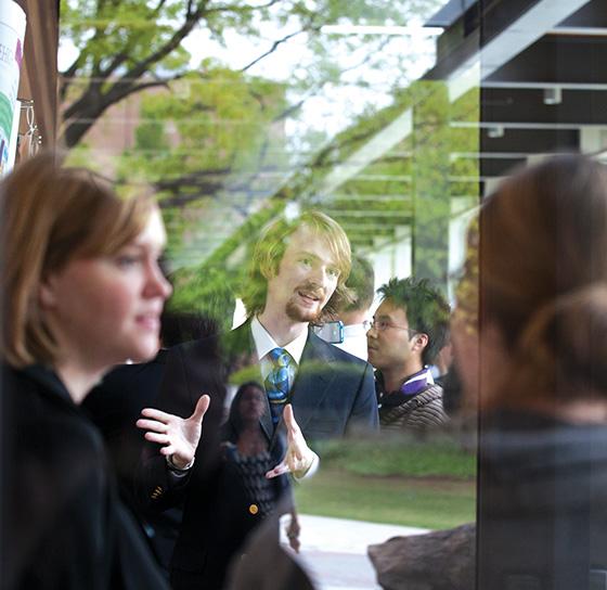 A group of people are engaged in a lively conversation outside. The setting appears to be a casual, outdoor social gathering near some greenery. The individuals are dressed in semi-formal attire and are smiling and gesturing expressively.