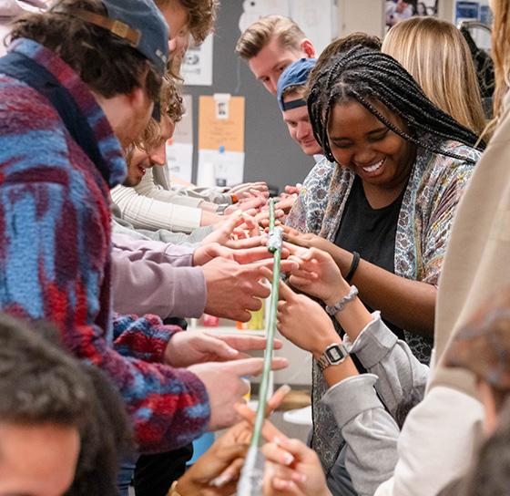 A group of people gather around a long table, focused on constructing tall structures with what appear to be sticks or thin rods. They seem engaged and collaborative, with one woman in the center smiling as she works on the project.