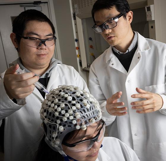 Three people are in a lab-like setting. Two individuals stand and wear white lab coats and glasses. They are looking at and discussing with a seated person who is wearing a cap covered in electrodes, possibly for a medical or scientific experiment.