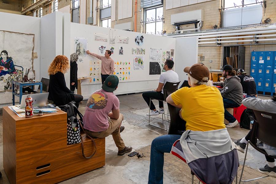 A presenter is standing at the front of a room pointing at a whiteboard, while a group of people sit and listen attentively. The setting appears to be a casual workshop or an educational session, with various notes and images displayed on the walls around the presenter.