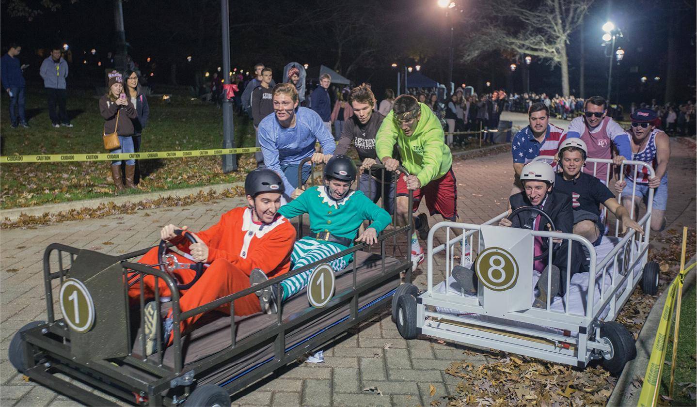 People dressed in various costumes, including a Santa outfit and a green elf, are participating in a race with homemade soapbox cars resembling beds. Spectators watch from behind barricades at nighttime, and the area is well-lit with artificial lights.