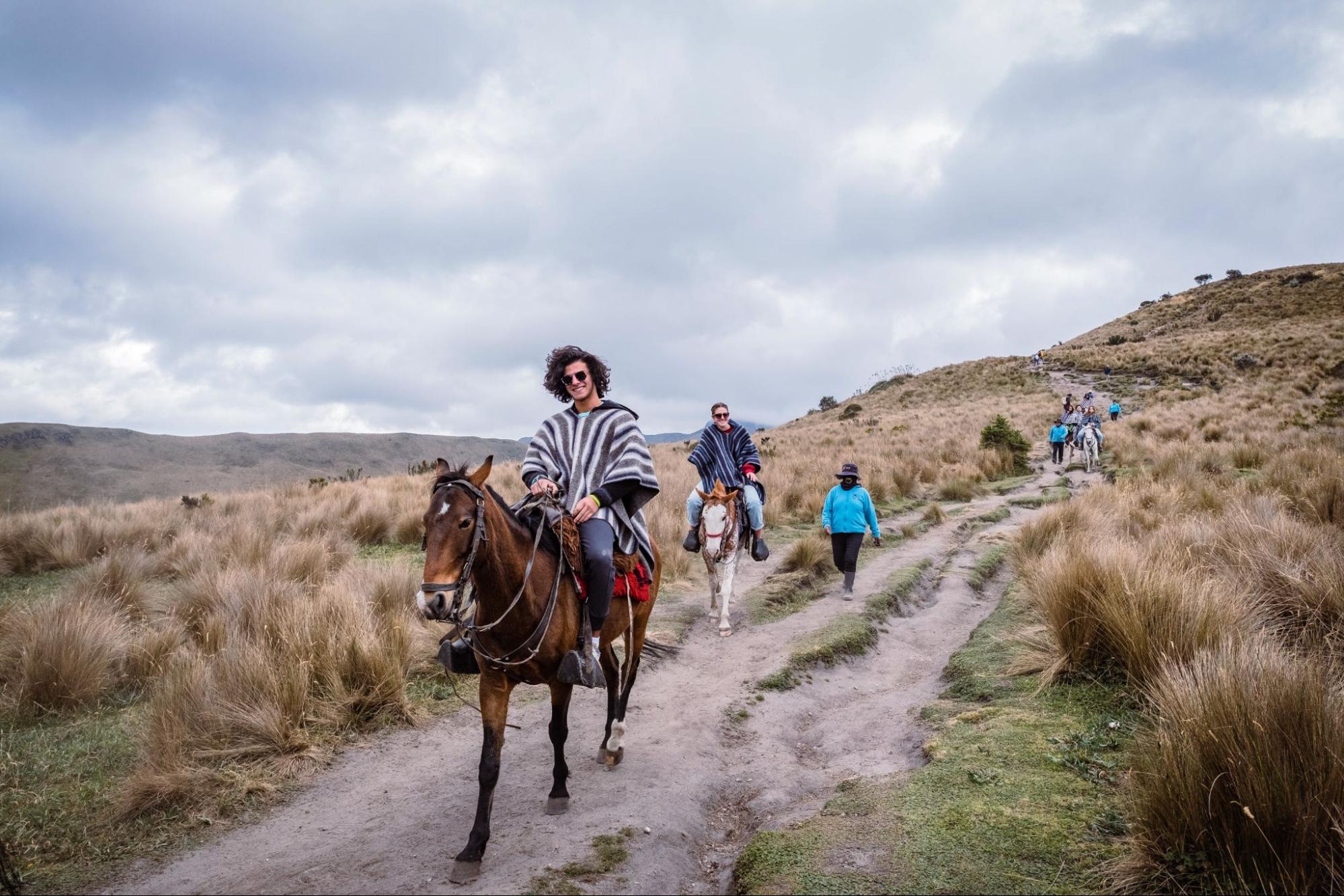 A group of people, including a person on horseback, travels along a grassy dirt path in a hilly landscape. The sky is overcast, and the terrain is covered with patches of grass and scattered shrubs.