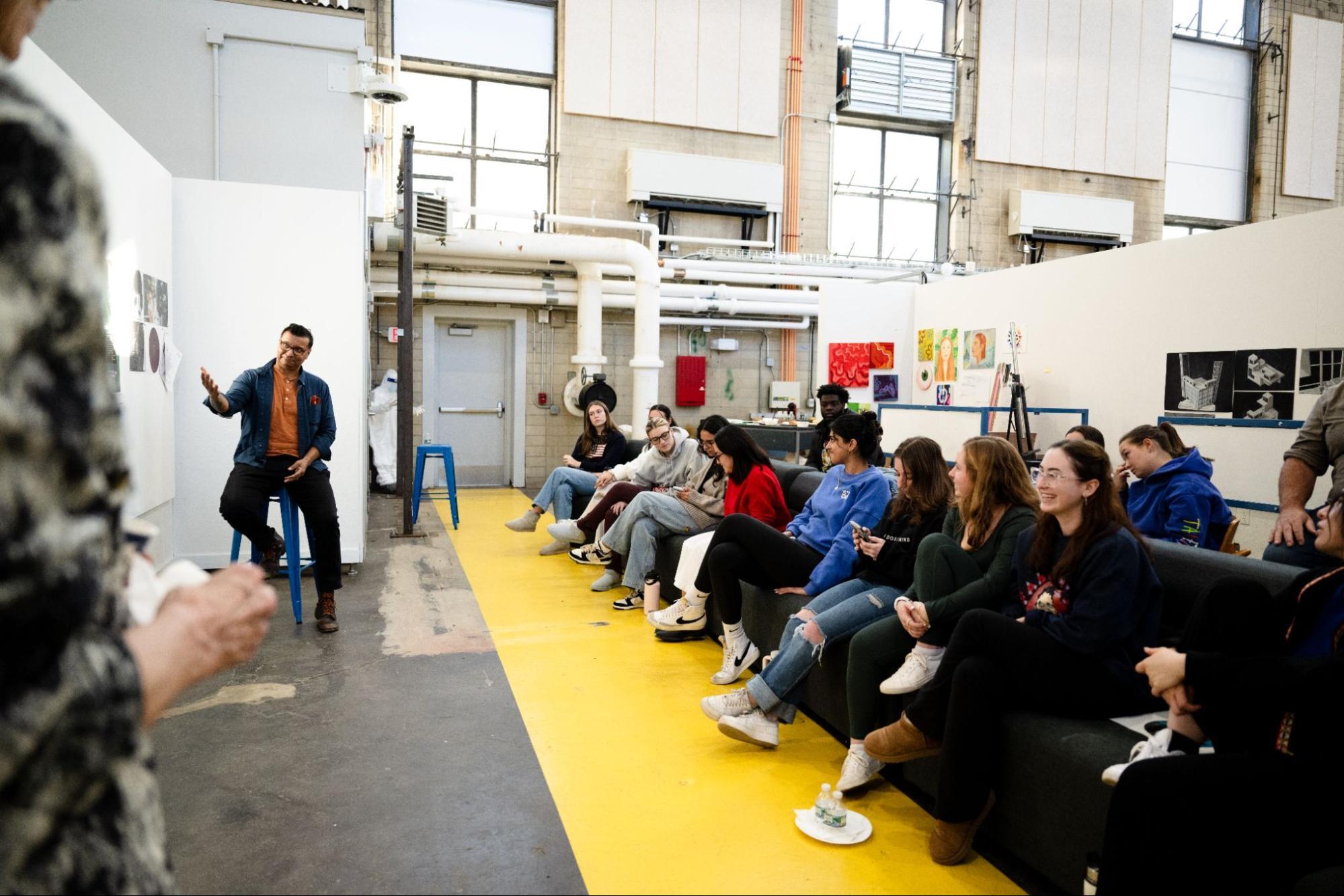 A group of people sit attentively on chairs arranged in a line along a bright yellow floor. A person stands in front of them gesturing, possibly giving a presentation or leading a discussion. The setting appears to be an open, well-lit indoor space.
