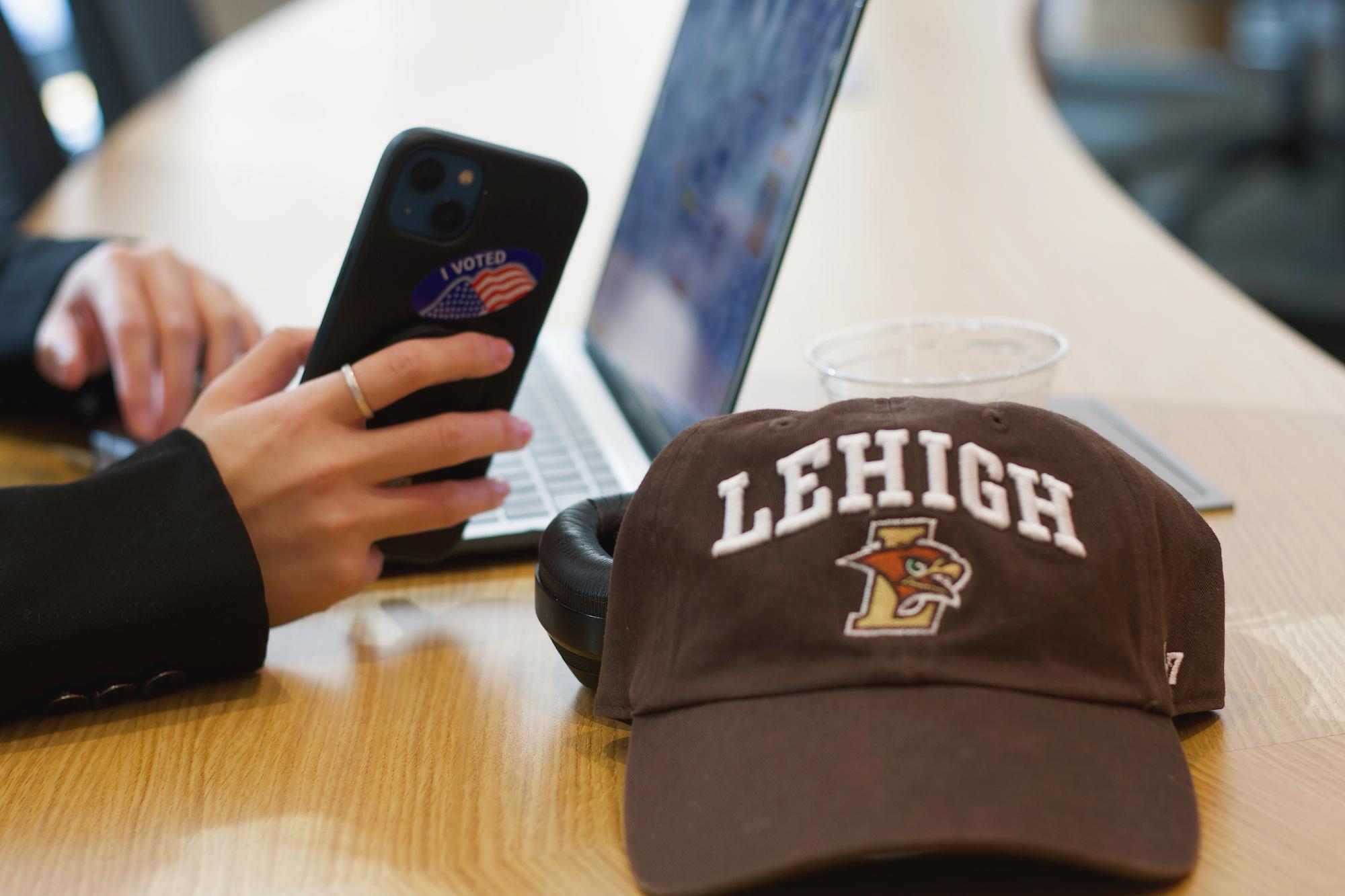 A Lehigh baseball cap sit on a table in front of a student holding a phone with an "I voted" sticker