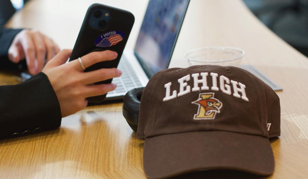 A Lehigh baseball cap sits on a table in front of a student holding a phone with an "I voted" sticker on it.