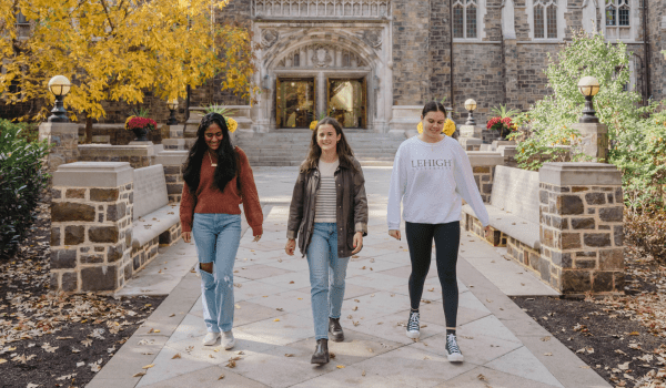 Three people walk along a tree-lined path on the Lehigh University campus. They're wearing casual clothes as they head towards a stone building. The scene is bright and autumnal.