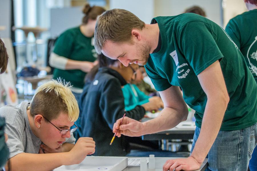 A male Lehigh student in a green shirt assists a younger student with a writing task. Other students are engaged in similar activities in the background. The setting appears to be a classroom.