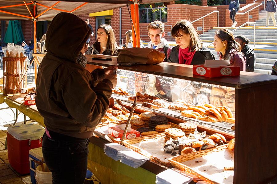 A person in a hoodie photographs a vibrant food stall at a market. Various baked goods and pastries are displayed, while vendors behind the counter smile and engage with customers.