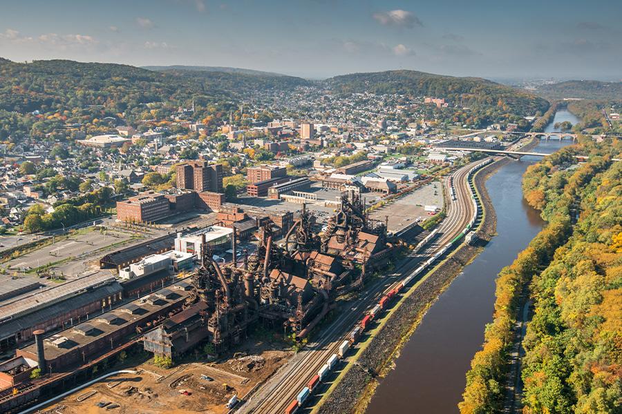 Aerial view of Bethlehem with the Lehigh River running alongside a railway and roads. Buildings and industrial structures are scattered throughout, with hills in the background.