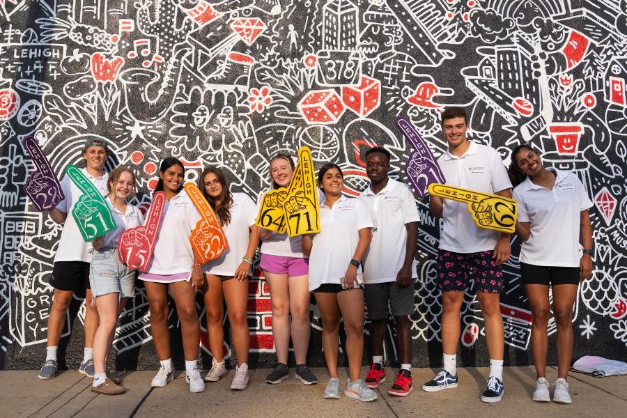 A group of people standing in front of a colorful, graffiti-covered wall. They are wearing casual white t-shirts and shorts, holding paint brushes and rollers. The scene has an artistic and playful atmosphere.