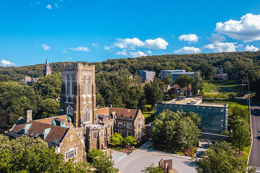 Aerial view of the Alumni Memorial Building, a historic stone building with a tall tower, surrounded by lush greenery and trees. In the background, more buildings and a partly cloudy blue sky are visible.