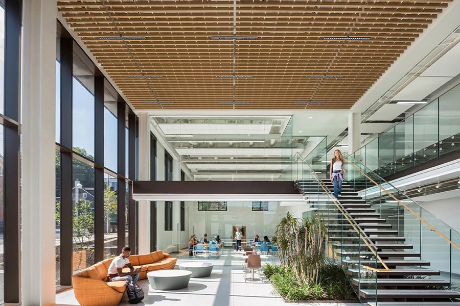 Interior of HST Building - modern and spacious with large windows letting in natural light. Features include a glass staircase, wooden ceiling panels, and vibrant orange seating areas. Green plants add a touch of nature to the sleek design.