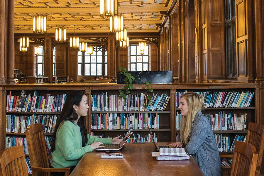 Two people sit at a wooden table in a large library with high ceilings and ornate light fixtures. They are surrounded by bookshelves filled with books. Large windows let in natural light.