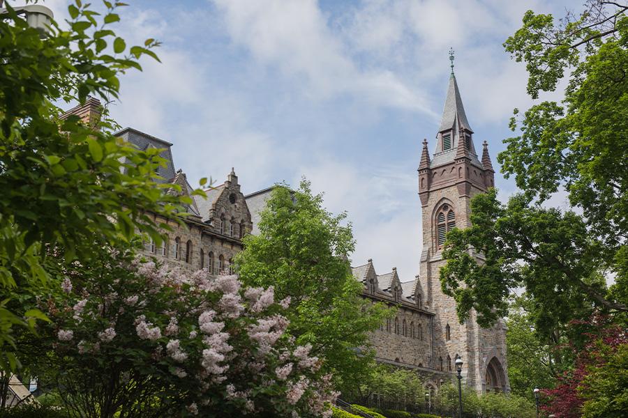 Clayton University Center - A historic university building with a tall clock tower stands against a partly cloudy sky. Lush green trees and blooming bushes frame the scene.