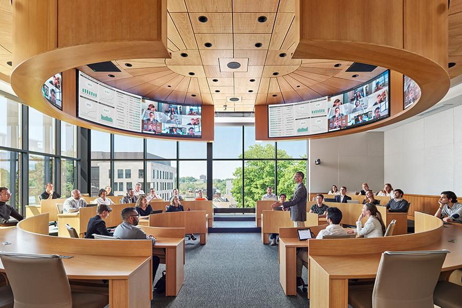 A modern classroom with large windows and circular wooden ceiling. Students sit at tables, engaging in a lecture. Multiple screen displays are mounted above, and sunlight filters through the glass, illuminating the space.