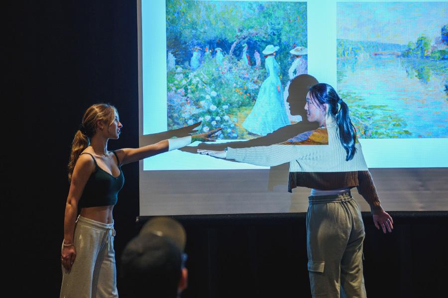 Two women stand in front of a screen displaying artwork. They are pointing towards each other, positioned to the right and left of the screen.