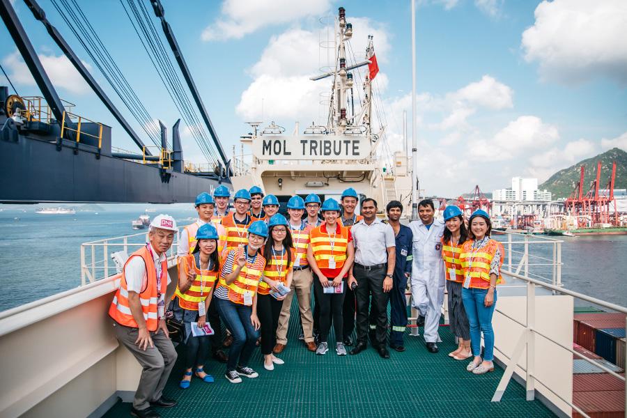 A group of people wearing safety vests and helmets pose for a photo on a ship's deck. They are standing in front of a large vessel with cranes, under a partly cloudy sky.