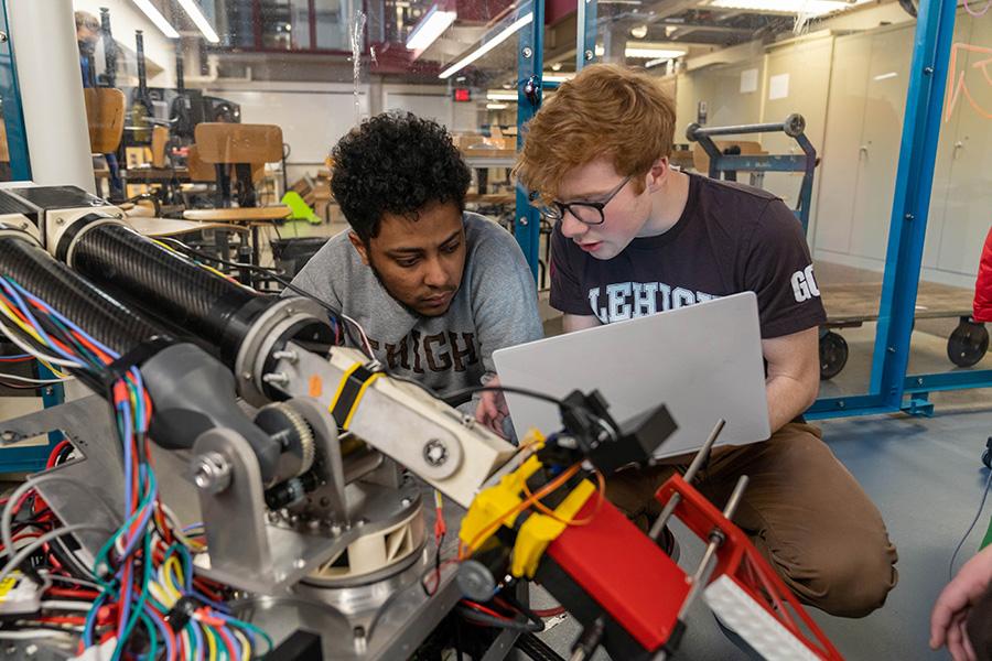 Two people working on a complex machinery setup in a workshop. They are focused on examining and adjusting components, with one holding a laptop. The background shows shelves and various tools.