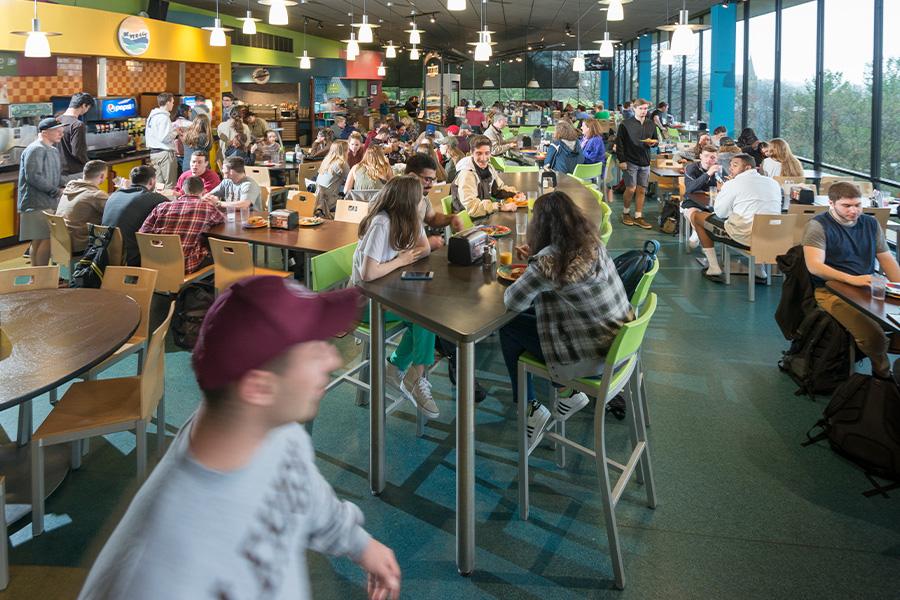 A bustling cafeteria filled with people sitting at tables, eating and talking. The space is well-lit with ceiling lights and large windows on the right. Foreground shows a person with a red cap, slightly blurred, walking past.