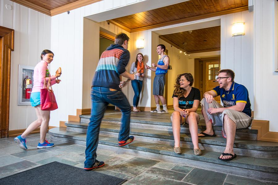 A group of young adults casually hang out indoors. Some are seated on stone steps, while others stand and converse near a wooden doorway. The atmosphere is relaxed and social.