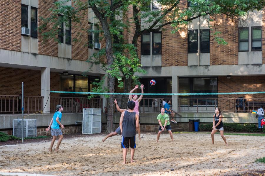 A group of people play volleyball on a sandy court surrounded by brick residence hall buildings and trees. One person is leaping to hit the ball over the net, while others watch and prepare for their next move.