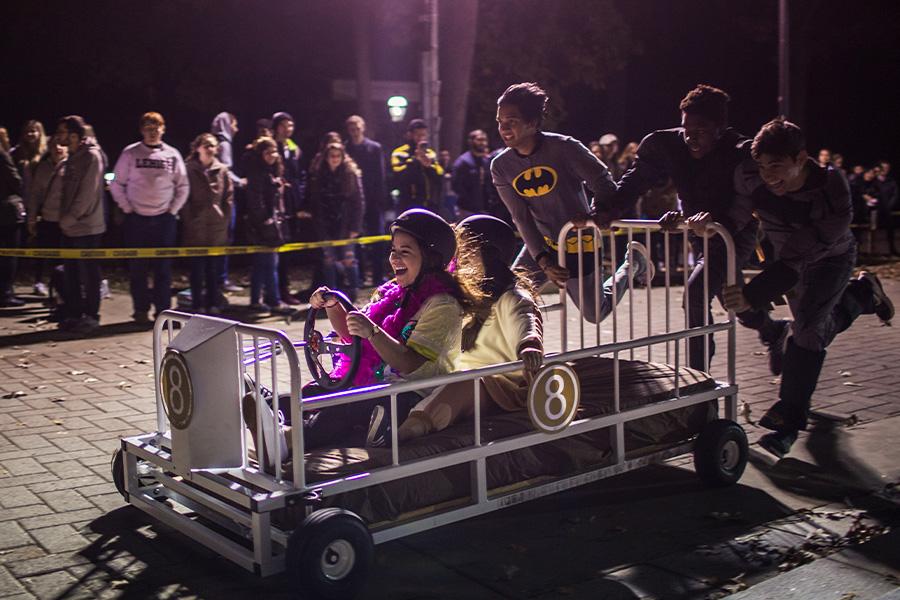 A group of people is participating in a nighttime event. Two children are seated in a handmade cart, resembling a bed, surrounded by adults. A crowd watches from the background, visible over a barrier.