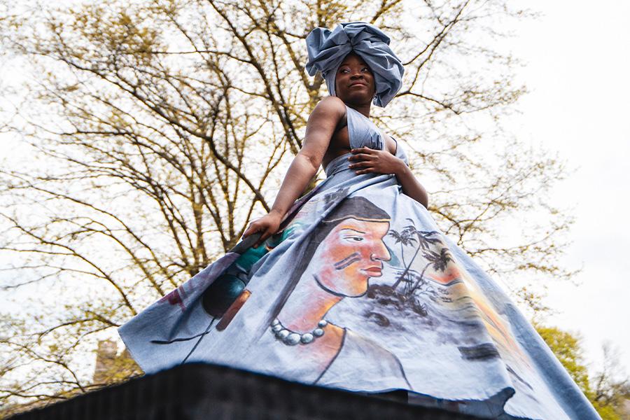 A woman wearing a long blue dress with a painting on the skirt, against a backdrop of trees with bare branches.