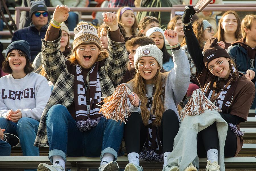 A group of people sitting on bleachers at the Lehigh Lafayette football game. They are wearing Lehigh clothes and hats, and some individuals have their arms raised, smiling and cheering.