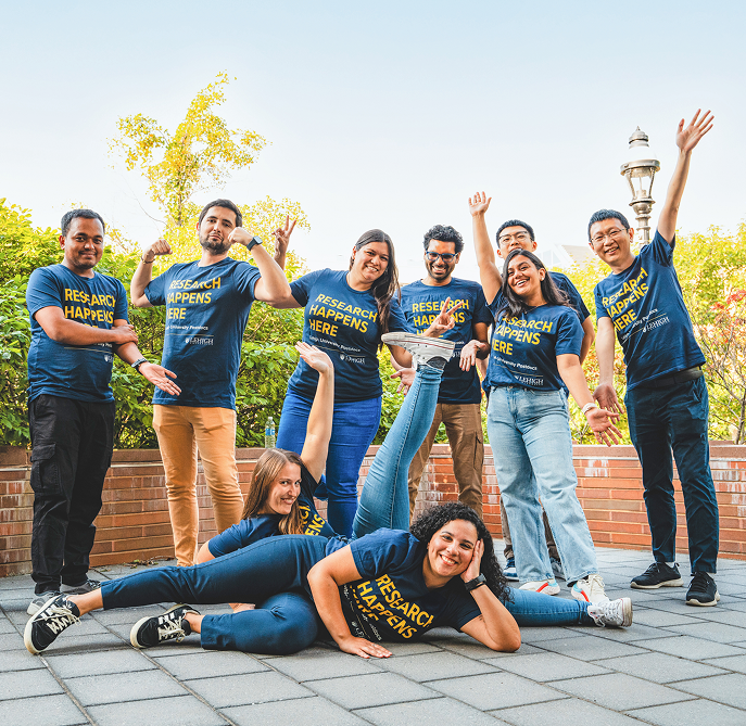 A group of nine smiling people in matching blue "Research Happens Here" shirts pose energetically outdoors; some stand with raised arms while others sit or lie on the ground playfully. Greenery and a lamppost are in the background.
