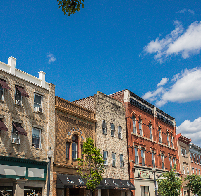 A row of historic brick buildings with shops and awnings under a bright blue sky with scattered clouds; a small tree stands in front of the storefronts.