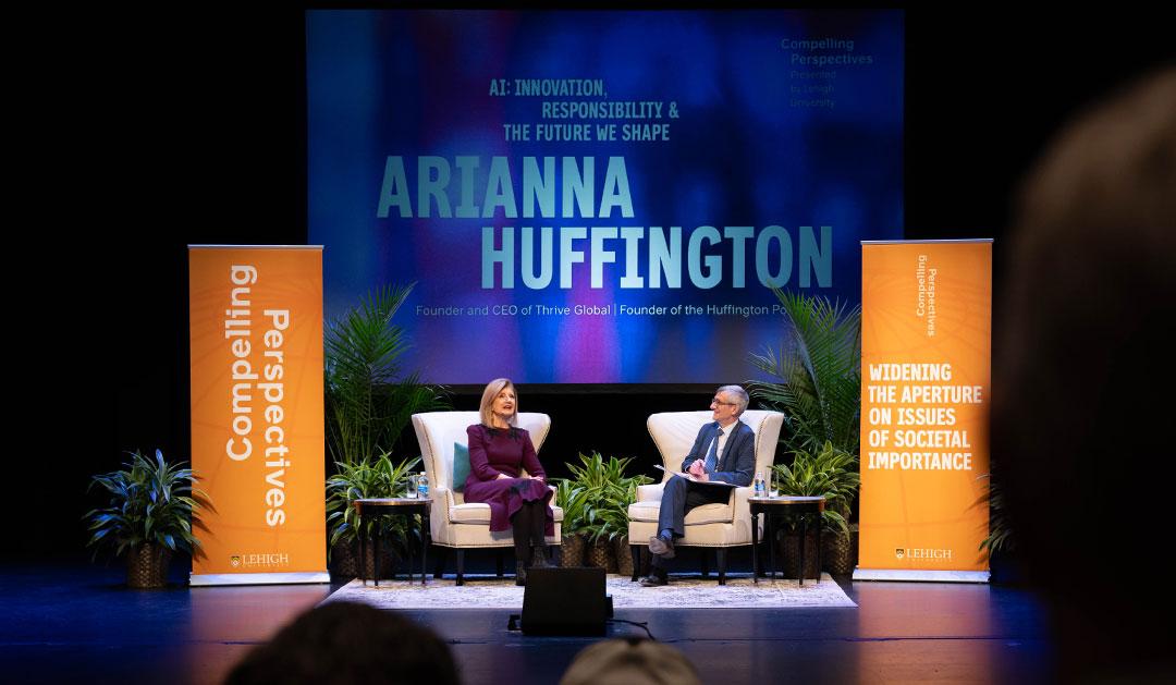 Ariana Huffington seated on stage, conversing with President Joseph Helble in a lit auditorium.