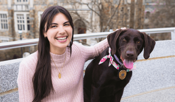 A woman with long dark hair wearing a light pink sweater smiles while sitting next to a chocolate Labrador wearing a pink collar and bandana. They are outdoors, with trees and buildings in the background.