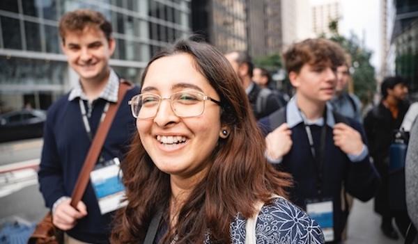 A young woman with glasses smiles at the camera while two young men with conference badges stand behind her on a city street. Tall glass buildings are visible in the background.
