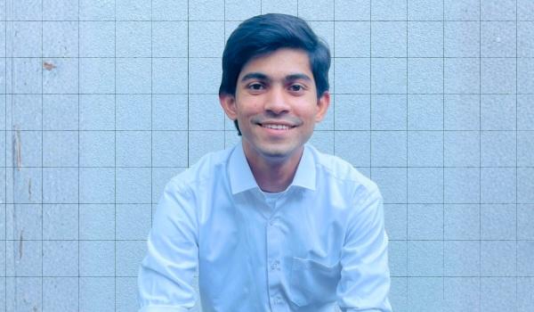 A young man with dark hair, wearing a light blue button-up shirt, sits and smiles in front of a white tiled wall.